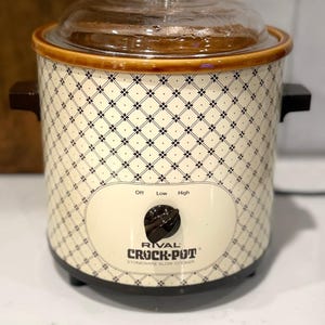 Puede incluir: Una olla de cocción lenta de gres Rival Crock-Pot con exterior color crema y un patrón de diamantes y flores negras. Tiene una tapa de vidrio transparente, dos asas laterales de color marrón oscuro y una perilla de control central negra con las marcas Off, Low, High.