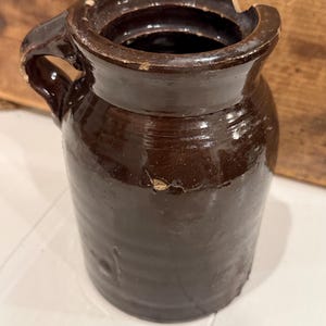 May include: A dark brown glazed ceramic crock with a single side handle and a cylindrical body. The rim features a noticeable chip, and there is a visible crack running vertically down the side of the vessel, showing signs of age and wear.