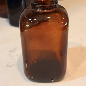 Puede incluir: Una pequeña botella de vidrio ámbar de forma rectangular con esquinas redondeadas y un cuello corto y estrecho. El vidrio es translúcido y marrón, con una superficie lisa y pulida, una abertura ancha en la parte superior y una base plana.