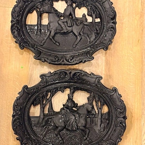 Può includere: Due placche da parete ovali nere con complessi rilievi. La placca superiore raffigura un cavaliere a cavallo in un paesaggio. La placca inferiore mostra una persona a cavallo in una zona boscosa con un edificio in lontananza. Entrambi i pezzi presentano bordi ornati e decorati.