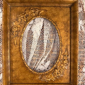 May include: A rectangular gold-toned picture frame with a central oval opening. The frame features raised floral relief detailing in the top-left and bottom-right corners, with a textured, weathered finish across the entire surface.