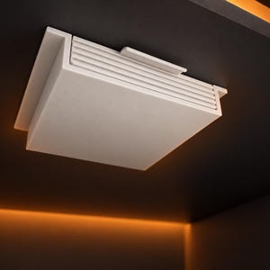Könnte beinhalten: Eine quadratische, weiße Deckenleuchte mit matter Oberfläche und einem gestuften, gerippten Design im oberen Bereich. Die Leuchte ist bündig an einer dunklen Fläche montiert und besitzt eine glatte, flache Unterseite sowie ein klares Profil.