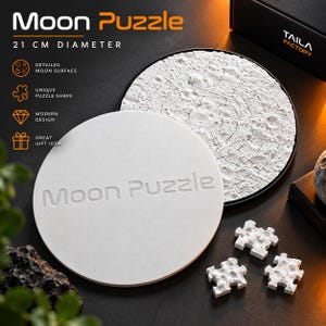 Könnte beinhalten: Ein kreisförmiges Moon Puzzle mit 21 cm Durchmesser und einer strukturierten weißen Oberfläche, die Mondkrater darstellt. Das Set enthält einen weißen, runden Deckel mit der Aufschrift Moon Puzzle, eine schwarze Box und drei Puzzleteile.