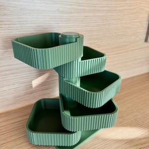 Peut inclure: Un organisateur de rangement à étages vert sauge composé de quatre plateaux carrés rotatifs fixés à un pilier central vertical. Chaque plateau présente une texture extérieure nervurée et une surface intérieure lisse, disposés en spirale.