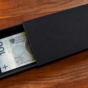 Könnte beinhalten: Eine schwarze, rechteckige Schiebeschachtel mit matter Oberfläche und einer kleinen Zuglasche an der Seite. Im offenen Fach liegt ein polnischer 100-Zloty-Geldschein mit dem Text Narodowy Bank Polski und der Zahl 100.