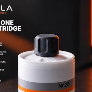 Pode incluir: Uma tampa de cartucho de silicone preta assenta sobre um cartucho cilíndrico branco rotulado como Nr.22. O texto TAILA FACTORY SILICONE CARTRIDGE CAP é visível, juntamente com ícones e texto indicando que mantém o conteúdo fresco e é reutilizável.