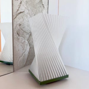 Puede incluir: Un florero blanco con un diseño vertical, plisado y retorcido. La estructura presenta una base cuadrada de color verde sólido. El objeto tiene una forma geométrica en espiral que se estrecha hacia el centro y se ensancha arriba y abajo.
