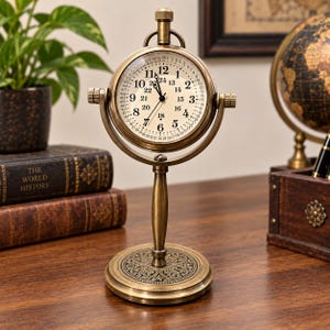 May include: A brass-toned metal desk clock featuring a circular face with both 12-hour and 24-hour markings, along with a secondary outer ring of numbers. The clock is mounted on a swivel frame with a pedestal base and a decorative filigree pattern.