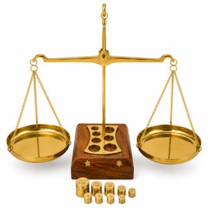 Puede incluir: Una balanza de metal dorado con dos platos colgantes suspendidos por cadenas de una viga central. La balanza cuenta con una base de madera con un soporte de pesas de latón y dos incrustaciones de estrellas. Diez pesas cilíndricas de latón.