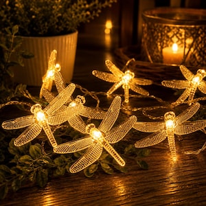May include: A string of decorative dragonfly-shaped lights featuring clear, textured wings and bodies that emit a warm, golden glow. Each dragonfly is connected by a thin wire, creating a series of illuminated insect figures with detailed wing patterns.