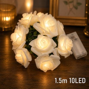 May include: A string of 10 LED lights shaped like white roses, emitting a warm glow. The set includes a clear plastic battery pack and measures 1.5 meters in length. The roses are interspersed with small green leaves along the thin wire cord.
