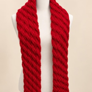 May include: A vibrant red knitted scarf featuring a prominent diagonal cable stitch pattern. The scarf is draped vertically, showcasing the thick, textured yarn construction and the consistent, repeating twisted design that runs the entire length.