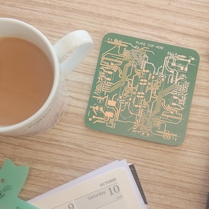 May include: A square green coaster featuring a gold-colored printed circuit board pattern and the text "PLACE CUP HERE" at the top. The coaster is positioned next to a white ceramic mug filled with a light brown beverage and a laptop computer.