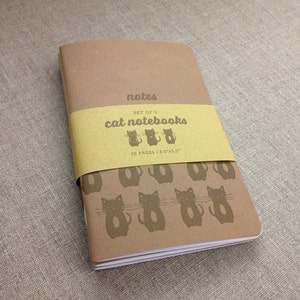 May include: Set of three brown cat notebooks with 36 pages each. The notebooks are 3.5 inches by 3.5 inches and feature a repeating pattern of black cats on the front cover.