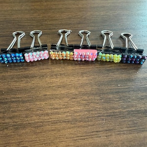 May include: Six black metal binder clips arranged in a row, each featuring a decorative front panel covered in small, multi-coloured iridescent rhinestones. The colours include blue, light pink, orange, bright pink, yellow, and dark purple.