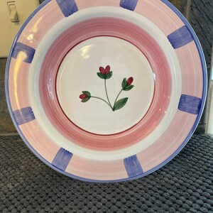 May include: A round ceramic bowl featuring a white center with a small hand-painted floral motif of three red buds on green stems. The wide rim is decorated with concentric pink bands and evenly spaced vertical blue brushstroke rectangles.