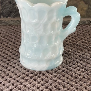 Puede incluir: Una jarra de vidrio azul claro con un patrón texturizado similar a burbujas. La jarra tiene un borde festoneado y un asa curvada. El vidrio tiene una cualidad lechosa y opalescente. Se encuentra sobre una superficie texturizada.
