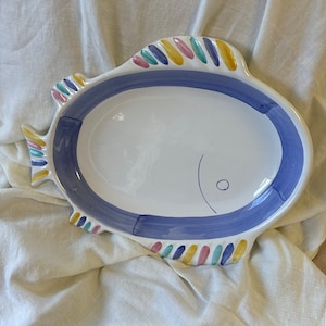 May include: A white ceramic serving platter shaped like a fish. The platter features a wide blue painted border around the center, with a simple line and circle detail for an eye. The fins and tail are decorated with small, colorful stripes.