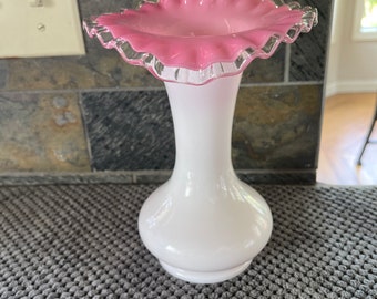 May include: A white glass vase with a ruffled, pink rim. The vase has a bulbous base and a slender neck. The rim is a vibrant pink color, and the vase is set against a neutral background.