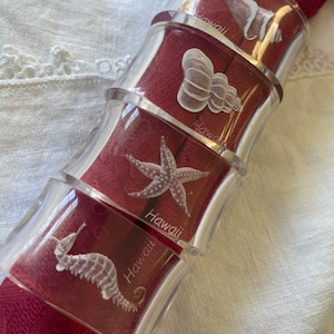 May include: Clear acrylic napkin rings with etched sea life designs, including a seahorse, starfish, and seashell. The word "Hawaii" is etched on each ring. The rings are wrapped around a red napkin, creating a decorative table setting.