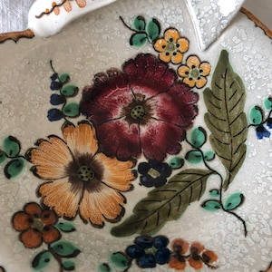 May include: A heart-shaped ceramic dish with a handle, featuring a floral design. The flowers are in shades of burgundy, orange, and yellow, with green leaves and blue accents. The rim is scalloped and has a brown trim.