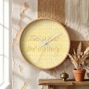 May include: A round wall clock with a light wooden frame and a yellow patterned face. The clock features white hour and minute hands and cursive text in the center that reads, Time is now the currency, with white lines marking the hour positions.