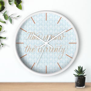 Op de afbeelding: Een ronde wandklok met een witte rand en een lichtblauwe geometrische achtergrond. De wijzerplaat bevat de koperkleurige cursieve tekst Time is now the currency en is voorzien van dunne koperen uurmarkeringen en witte wijzers.