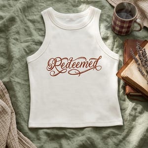 Op de afbeelding: Een wit geribbeld hemdje met het woord "Redeemed" in een donkerbruin, gestileerd cursief lettertype op het midden van de borst. Het kledingstuk heeft een hoge, ronde halslijn en een mouwloos, nauwsluitend ontwerp.