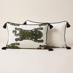 May include: Two rectangular throw pillows featuring a white base with a repeating dark green tiger pattern. Each pillow is finished with black piping along the edges and black tassels at all four corners, providing a structured and decorative look.