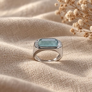 May include: A silver-toned ring featuring a rectangular, smooth, pale green stone set horizontally. The metal band is accented with small, clear, sparkling stones on both sides of the central setting, creating a polished and refined appearance.