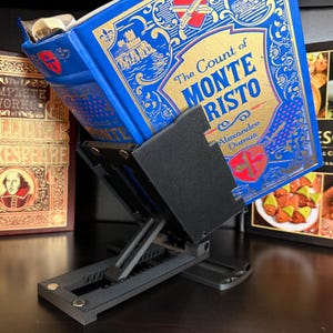 May include: A black adjustable book stand holding a blue hardcover copy of The Count of Monte Cristo by Alexandre Dumas. The stand features a flat base with a notched support arm and a rectangular panel that secures the open book in place.