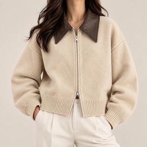 May include: A beige knit zip-up jacket featuring a contrasting dark brown pointed collar, long sleeves with ribbed cuffs, a ribbed hem, and a full-length silver-toned metal zipper closure down the center front of the garment.
