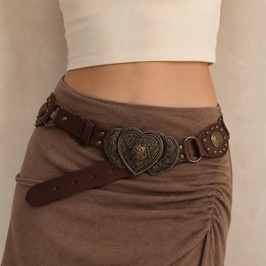 May include: A dark brown leather belt featuring a prominent central buckle shaped like a heart with intricate embossed detailing. The belt includes additional circular metal accents and hardware, worn over a brown ruched skirt and a white top.