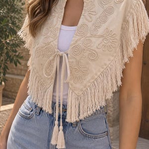 Peut inclure: Un gilet sans manches beige orné de broderies ton sur ton avec des motifs floraux en spirale, un décolleté en V et de longues franges sur les bords. Le vêtement comprend une fermeture à nouer sur le devant avec deux pompons suspendus.