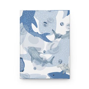 May include: A hardcover journal featuring a repeating pattern of various sharks and sea creatures in shades of blue and white. The design includes whale sharks and hammerhead sharks illustrated in a flat, graphic style against a white background.