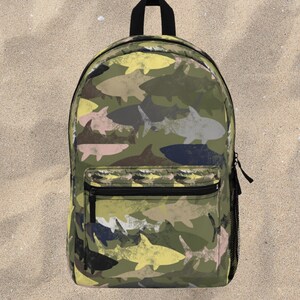 Puede incluir: Una mochila con un estampado integral de siluetas de tiburones en tonos verde oliva, amarillo pálido, azul oscuro y rosa suave. La bolsa incluye un asa superior negra, un bolsillo frontal con cremallera y un bolsillo lateral de malla negra.