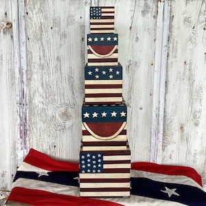 Puede incluir: Una pila vertical de cinco cajas cuadradas de madera con un tema patriótico desgastado en rojo, blanco y azul. Las cajas muestran patrones de bandera estadounidense, estrellas blancas sobre campos azules y rayas rojas y blancas.