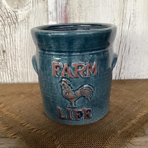 May include: A teal ceramic crock featuring two side handles and a raised relief design. The text "FARM" appears above a rooster illustration, with the word "LIFE" below. The glaze has a glossy finish with subtle variations in color depth.