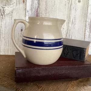 May include: A cream-colored ceramic pitcher featuring a prominent dark blue horizontal stripe pattern around its rounded body, a curved handle on the side, and a small pouring spout at the top rim, presented as a functional kitchen vessel.
