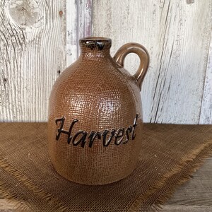 Peut inclure: Une cruche en céramique brune avec une surface texturée rappelant la toile de jute. La cruche présente une anse unique et un goulot étroit. Le mot Harvest est écrit en lettres cursives brun foncé sur le corps arrondi du récipient.