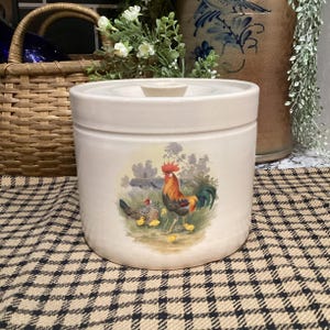 May include: A cylindrical off-white ceramic canister with a matching lid. The front features a colorful illustration of a rooster, a hen, and several small yellow chicks in a grassy field. The canister has a smooth, matte finish and a knob handle.