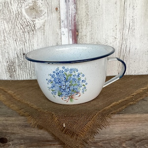 May include: A white enamel bowl with a speckled finish and a dark blue rim. The bowl features a floral decal of blue forget-me-not flowers tied with a light pink ribbon on the front. It includes a single side handle made of matching white enamel.