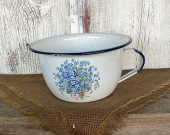 Op de afbeelding: Een witte emaille kom met een gespikkelde afwerking en een donkerblauwe rand. De kom is aan de voorzijde voorzien van een bloemendecoratie van blauwe vergeet-mij-nietjes, samengebonden met een lichtroze lint. Inclusief één zijhandvat van emaille.