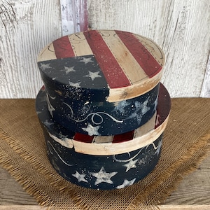 May include: Two stacked round wooden boxes featuring a distressed patriotic design. The lids and sides are painted with navy blue, red, and cream stripes and stars, with the text God Bless America written in cursive script on the top lid surface.