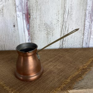 May include: A copper coffee pot with a wide, rounded base, a narrow neck, and a flared rim. It features a long, slender, metallic handle attached to the side. The exterior has a warm, metallic finish with visible signs of use and natural patina.