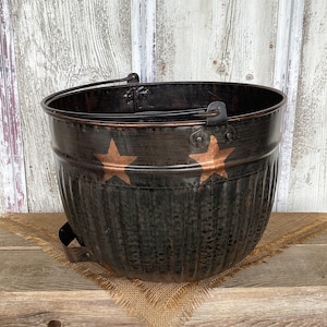May include: A dark brown, ribbed metal bucket featuring a copper-colored star cutout on the front. The bucket has a rounded shape, a wide open top, and a sturdy metal handle attached to the sides with riveted brackets for carrying or hanging.