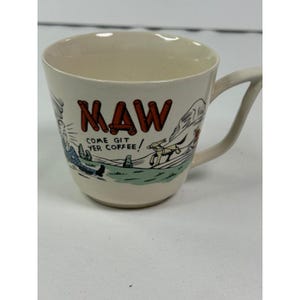Puede incluir: Una taza de cerámica de color crema con una ilustración gráfica de una persona y un perro en un paisaje. El texto "MAW" está impreso en letras rojas grandes sobre la frase "COME GIT YER COFFEE!" en negro. La taza tiene un asa lateral.