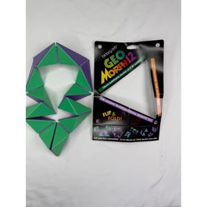 May include: A purple and green geometric puzzle toy composed of connected triangular segments, positioned next to its original black cardboard packaging. The packaging features the text "Binary Arts," "Geo Morph 12," "Create surprising shapes as if by magic!," "Challenge your imagination!," "The amazing transformational geometry toy!," and "Flip & Fold!."