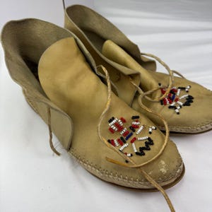 May include: A pair of tan leather moccasins featuring a high ankle cuff and a wrap-around leather lace tie. The top of each moccasin is decorated with a geometric pattern of small white, red, blue, and black seed beads, with visible stitching.