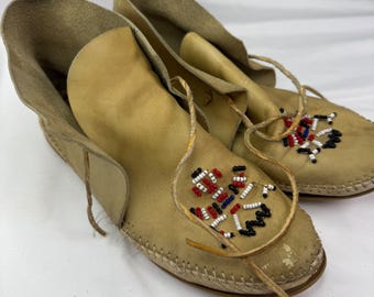 May include: A pair of tan leather moccasins featuring a high ankle cuff and a wrap-around leather lace tie. The top of each moccasin is decorated with a geometric pattern of small white, red, blue, and black seed beads, with visible stitching.